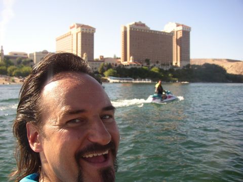 1990 Ford Bronco - smiling man takes a selfie on the river while a jet ski rider trails behind, with tall casino-resort towers beyond.