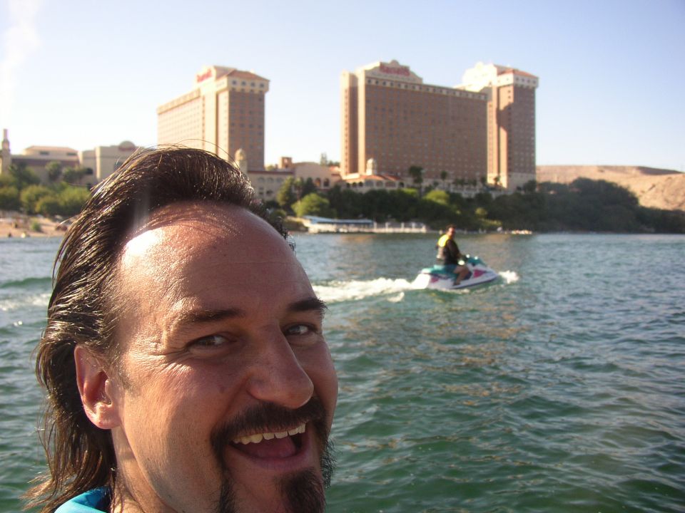 Selfie of a smiling man on the water with a jet ski rider and tall resort towers in the background