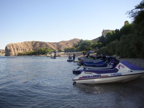 1990 Ford Bronco - line of purple-and-teal Sea-Doo jet skis sits beached along a riverbank below desert hills and a hilltop building.