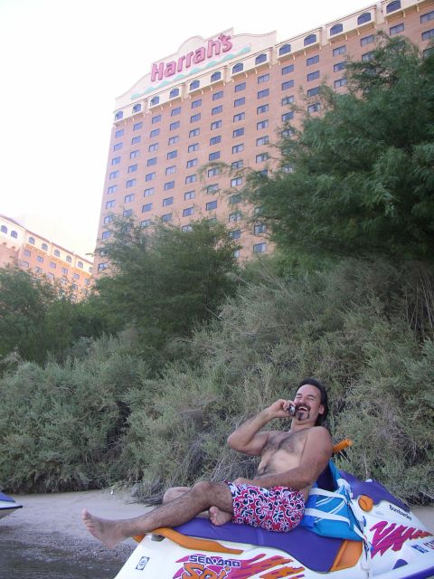 1990 Ford Bronco - shirtless man on a Sea-Doo jet ski talks on a phone amid riverside brush below a tall Harrah's casino hotel tower.