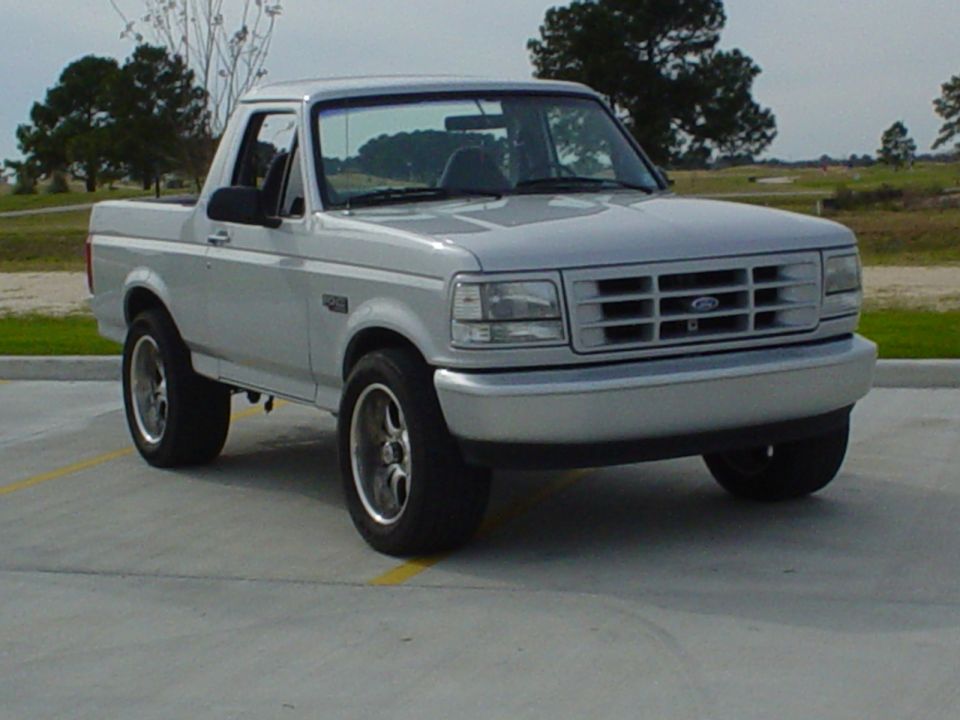 Silver Ford Bronco with top removed, front three-quarter view, aftermarket wheels, parked on pavement