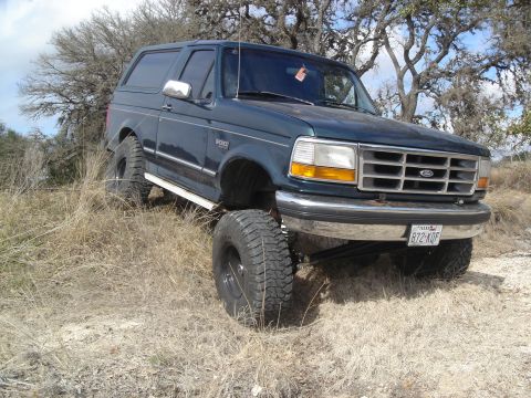 1995 Ford Bronco - This lifted Ford Bronco is shown off-road on a dry, grassy incline under a partly cloudy sky.