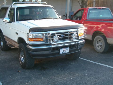 1996 Ford Bronco - A white Ford Bronco is parked next to a red pickup truck, showing its front grille and lights.