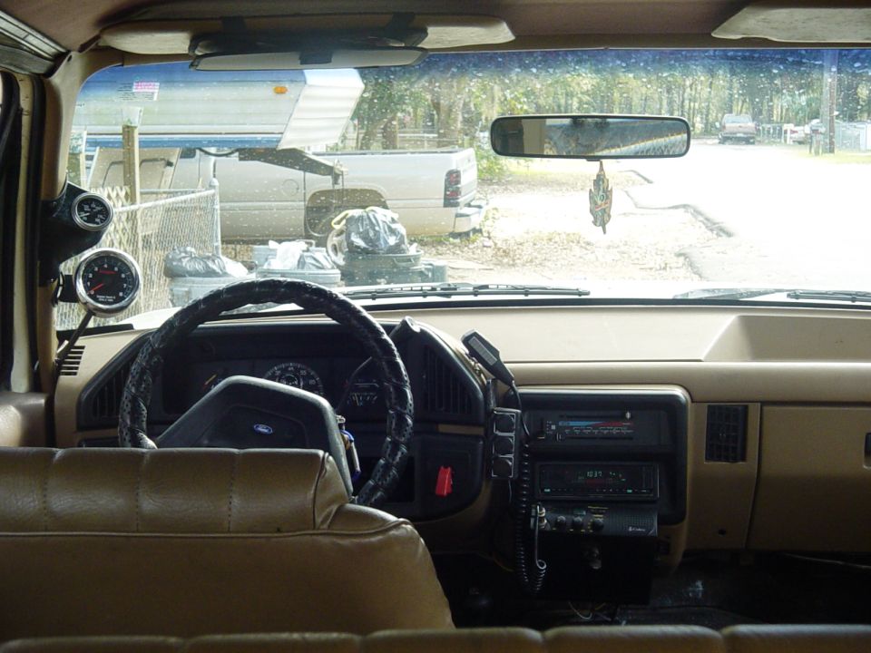 Tan Ford truck interior with steering wheel, aftermarket gauges and radio, junkyard out windshield