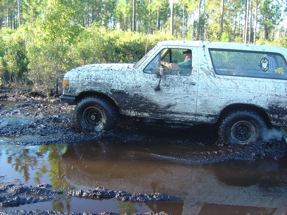 White SUV stuck in mud, driver visible. Looks like a 1987-1991 Ford Bronco