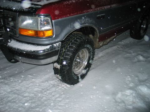 1995 Ford Bronco - Nighttime close-up of the front driver-side corner of a dark red and gray Ford Bronco/F-series truck in falling snow, showing the headlight…