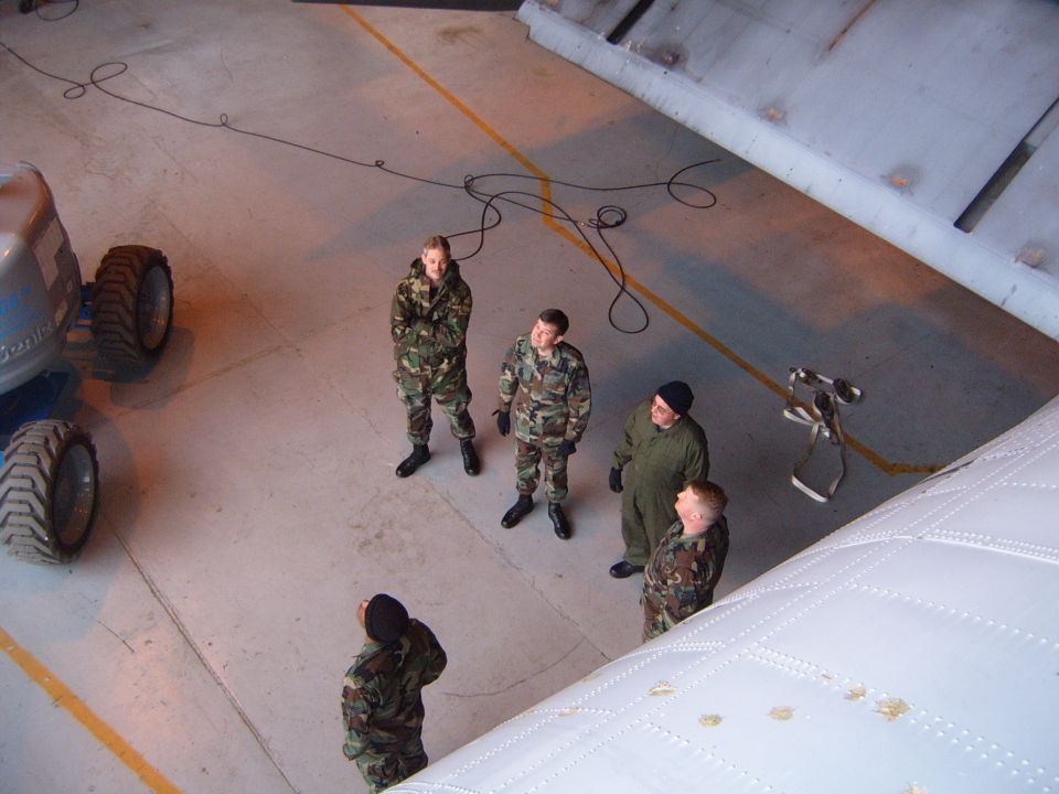 People in camouflage uniforms stand on a concrete floor near a large piece of machinery with big tires