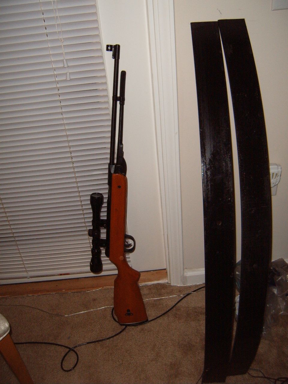 Air rifle with scope next to two black leaf springs, leaning against a wall with blinds
