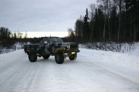 2003 Jeep TJ - A lifted pickup truck with camouflage paint drives on a snow-covered road through a wooded area.