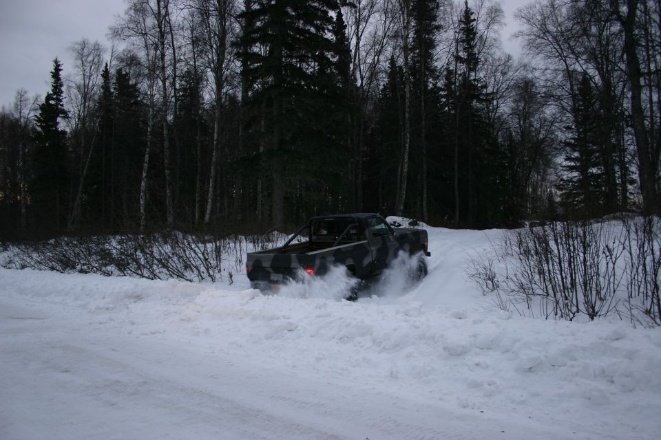 Pickup truck with camouflage paint and roll bar driving through deep snow, kicking up spray