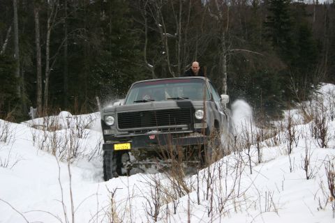 2003 Jeep TJ - A gray pickup truck navigates a snowy path, kicking up snow as it drives through a wooded area.