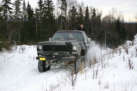 2003 Jeep TJ - A pickup truck with a camouflage paint job drives through a snowy landscape, spraying snow as it goes.