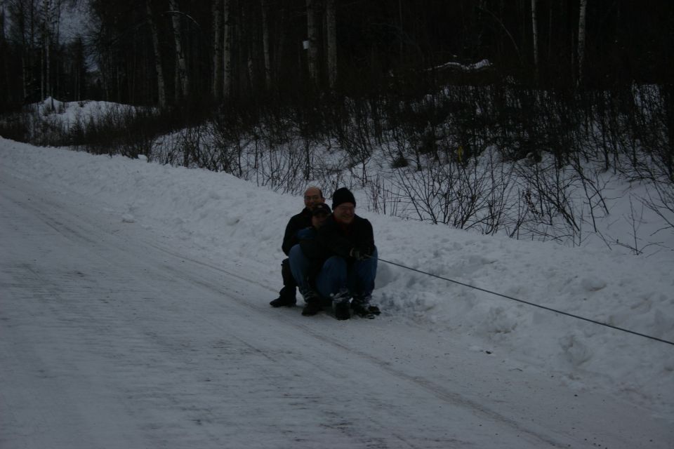 Two people on a sled being pulled up a snowy hill by a rope, with trees in the background