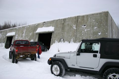 2003 Jeep TJ - A white Jeep Wrangler and a red pickup truck are parked in the snow near a large concrete building with open doorways.