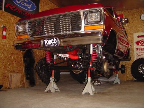 1978 Ford Bronco - The front end of a red pickup truck is elevated on jack stands, revealing its suspension and chassis.