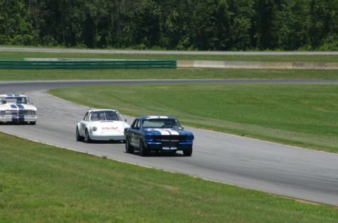 1965 Ford Mustang - Three race cars, including a 1965 Ford Mustang, compete on a paved track with grassy verges.