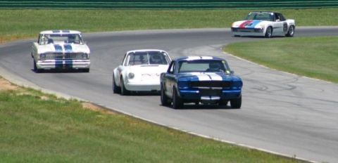 1965 Ford Mustang - Multiple vintage race cars compete on a paved track during a sunny day.
