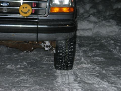1995 Ford Bronco - Low night shot of the front left corner of a dark Ford truck parked in packed snow, showing the grille with Ford oval, amber-lens headlamp,…