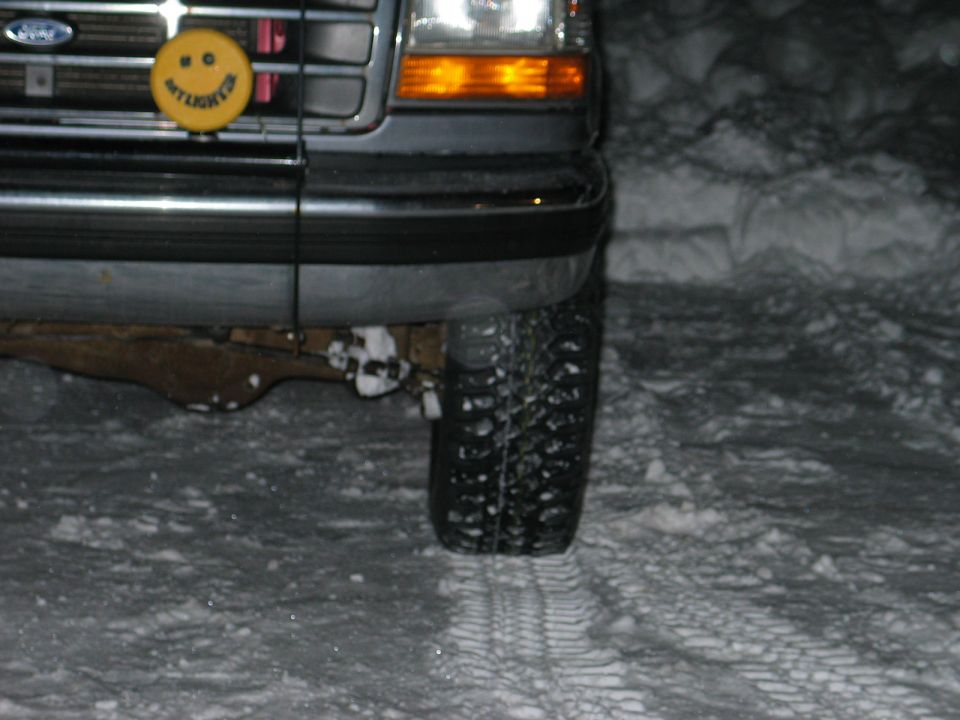 Front left corner of dark Ford truck in snow at night with yellow smiley light cover behind grille