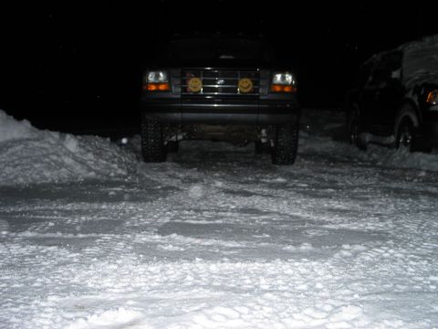 1995 Ford Bronco - Night shot of a lifted dark Ford 4x4 head-on in packed snow, parking lights glowing and two yellow smiley-face covers over the round drivin…