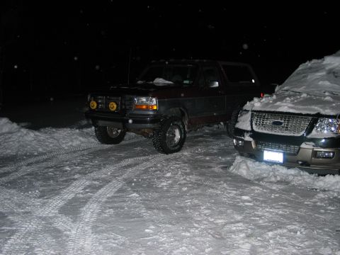 1995 Ford Bronco - Night shot in a snow-covered lot of a lifted two-tone dark red and gray Ford Bronco with a removable hardtop, seen from the 3/4 front drive…