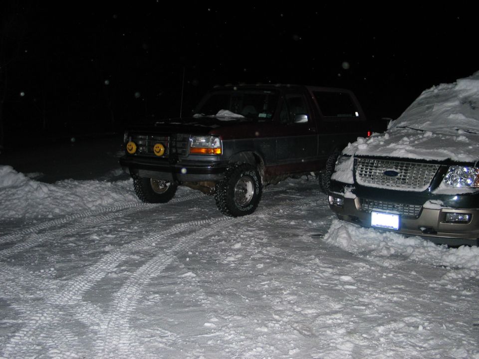 Lifted red and gray Ford Bronco with brush guard parked in snow at night, 3/4 front view