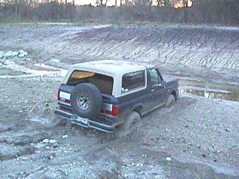 1989 Ford Bronco - A dark blue Ford Bronco with a tan top is stuck in a muddy, uneven terrain.