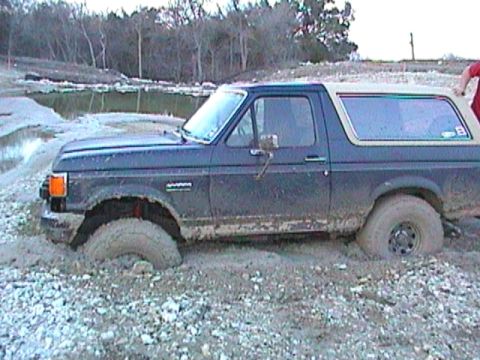 1989 Ford Bronco - A dark blue Ford Bronco is stuck in a muddy, rocky area with trees in the background.