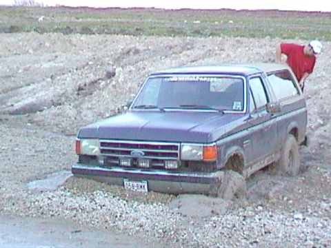 1989 Ford Bronco - A dark blue Ford Bronco is stuck in a muddy area with a person leaning over its roof.