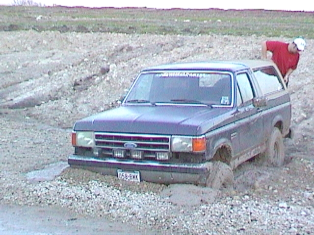 Dark blue Ford Bronco stuck in mud, front driver's side wheel submerged, person in red shirt leaning over roof