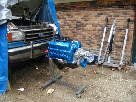 1990 Ford Bronco - A freshly rebuilt blue engine sits on a stand next to a blue truck, possibly undergoing maintenance.