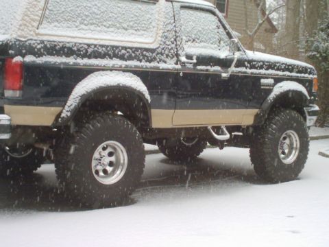 1989 Ford Bronco - A lifted Ford Bronco sits in the snow, its dark blue and tan paint contrasting with the white winter landscape.