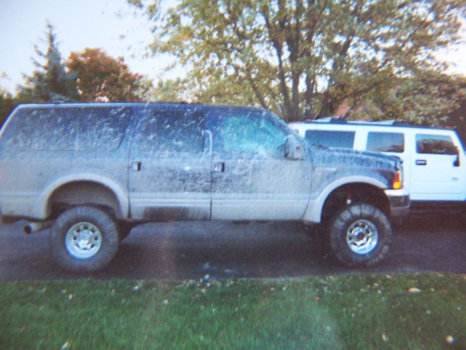 Mud-covered lifted dark blue Ford Excursion on big tires, parked beside a white Hummer H2