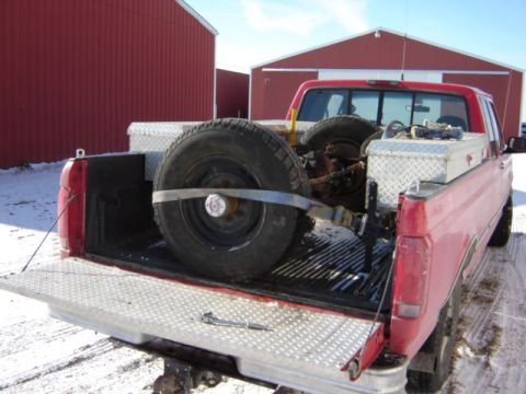 1992 Ford Bronco - Spare tire and axle are strapped into the bed of a red pickup truck, with red barns in the background.