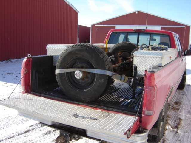 Red pickup truck bed with spare tire and axle secured by straps