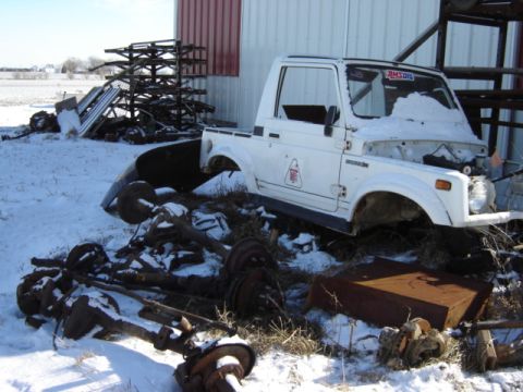 1992 Ford Bronco - A white Suzuki Samurai pickup truck is parked in a snowy yard with spare axles scattered nearby.