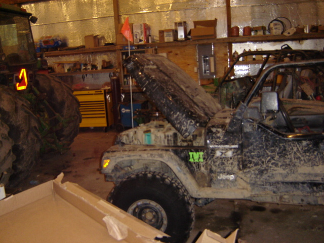 Mud-covered Jeep Wrangler with hood open, parked next to a tractor in a garage. Jeep has 'Wrangler X' and 'Jeep' badges