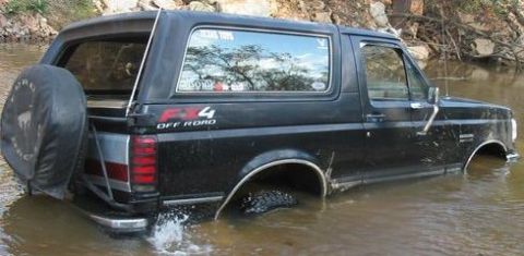 1988 Ford Bronco - A black Ford Bronco is stuck in shallow water, with its tires submerged and water splashing.