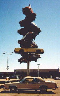 1985 Ford LTD Crown Victoria - A sculpture made of stacked cars stands tall against a blue sky, with a single car parked in front of it.