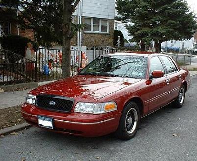 1999 Ford Crown Victoria - This red Ford Crown Victoria sedan is parked on a street in front of a house. The view is from the front three-quarter angle.