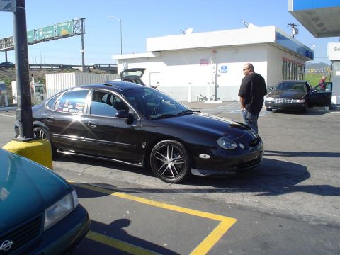 1993 Ford Taurus - This black sedan, likely from the early 2000s, is parked at a gas station with a second car visible at the pumps.