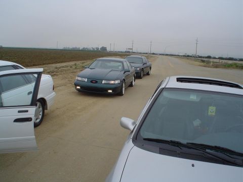 1993 Ford Taurus - A group of sedans are parked on a dirt road with fields in the background under an overcast sky.