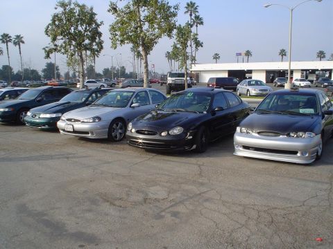 1993 Ford Taurus - Multiple sedans are parked in a lot under a clear sky, with trees and buildings in the background.