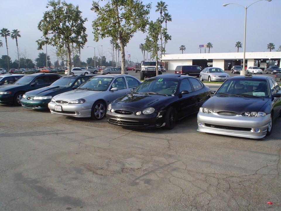 Several sedans parked in a lot, including a black car with a silver front bumper and a silver car with a black hood
