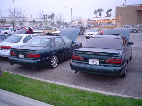 1993 Ford Taurus - Two dark green Ford Taurus sedans are parked side-by-side in a parking lot, one with its hood open.