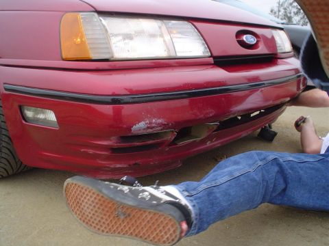 1993 Ford Taurus - A red 1993 Ford Taurus shows significant damage to its front bumper, with a person working on it.