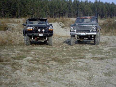 1976 Chevrolet C/K Pickup - Two modified pickup trucks are parked side-by-side on a dirt lot with trees in the background.