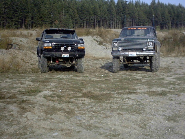 Two lifted dark 4x4 trucks parked front-facing on gravel near a pine forest.
