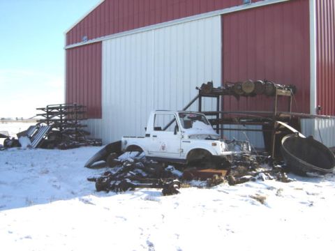 1992 Ford Bronco - A white Suzuki Samurai is partially visible in a snowy yard next to a large barn, surrounded by various vehicle parts.