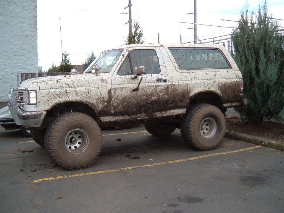 Mud-covered white lifted Ford Bronco with large off-road tires parked in a lot, side view